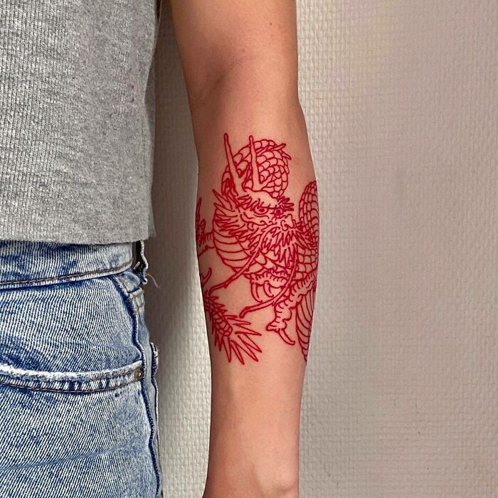Red ink tattoo of a detailed dragon design on forearm wearing a gray shirt and denim jeans.