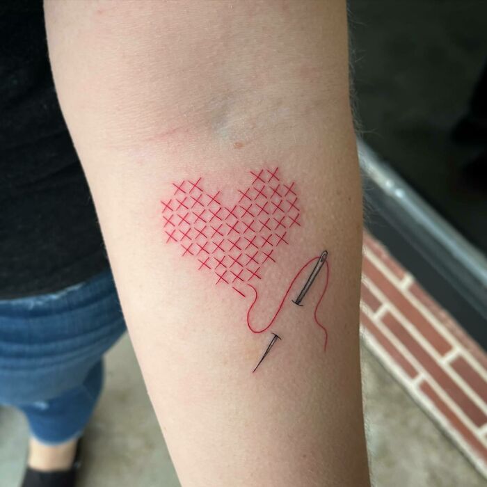 Red ink tattoo of a stitched heart with a needle on skin.