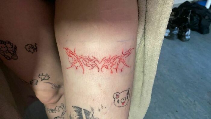 Red ink tattoo with intricate tribal design on the back of a leg, surrounded by small black ink tattoos.