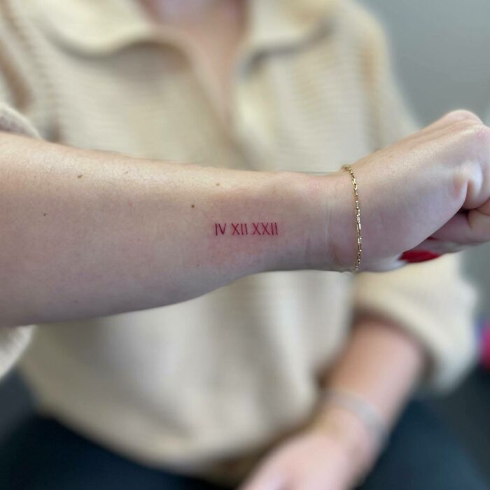 Red ink tattoo on forearm with Roman numerals IV XII XXII, close-up shot.