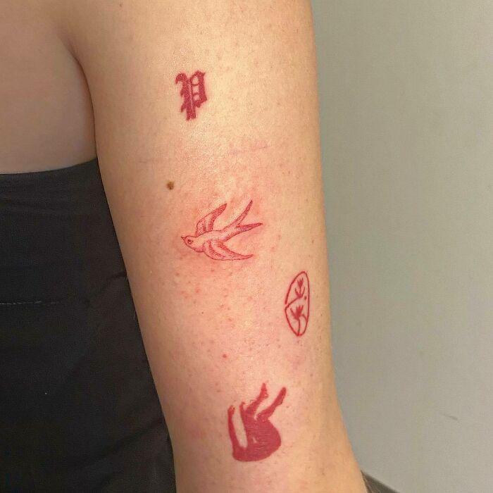 Red ink tattoos on an arm, featuring a stylized bird, letter, and abstract design.