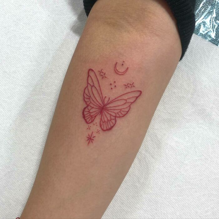 Red ink butterfly tattoo with crescent moon and stars on forearm.