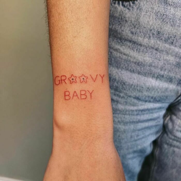 Red ink tattoo on forearm with the phrase "Groovy Baby" in a subtle design.