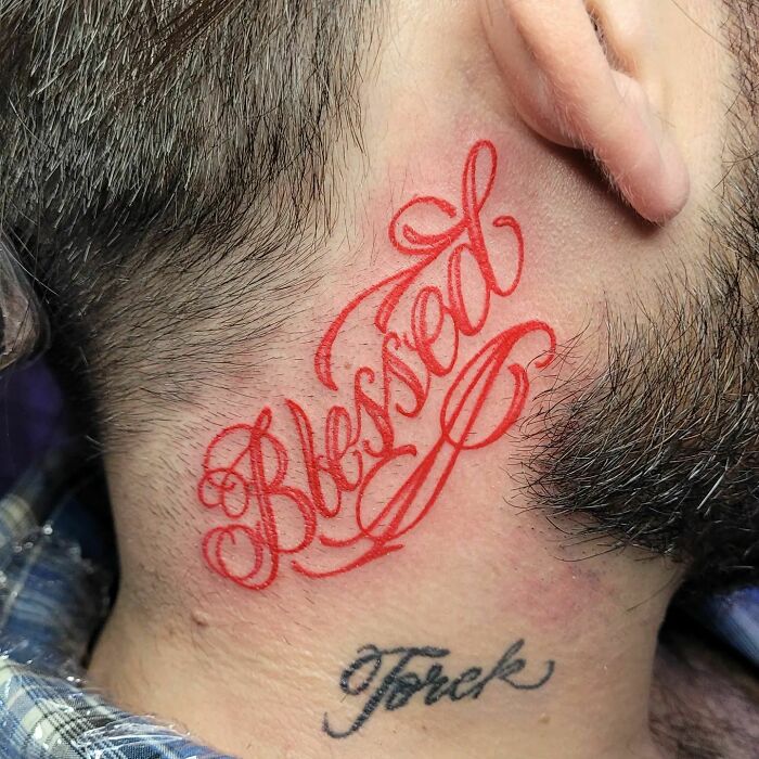 Red ink tattoo with "Blessed" text on the neck, showcasing intricate script design.