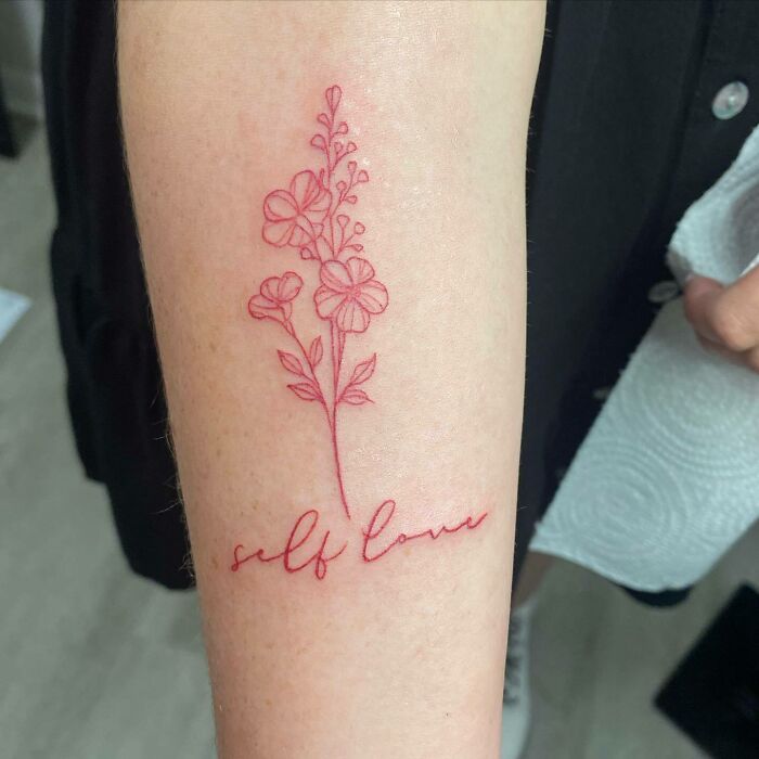 Red ink tattoo of flowers with "self love" text on forearm.