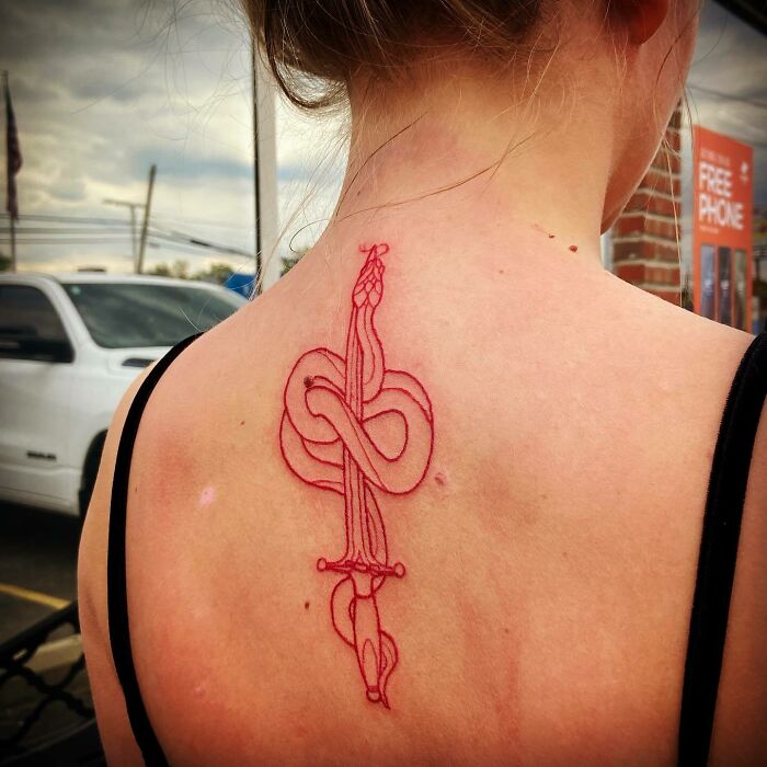 Red ink tattoo of a sword entwined with a snake on a person's upper back.