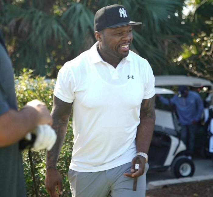 Celebrity playing golf, wearing a white shirt and black cap, outdoors near a golf cart.