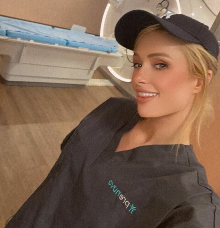 Celebrity in a work uniform and cap, smiling, in a room with medical equipment.