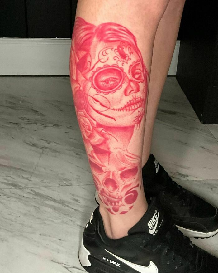 Red ink tattoo of a sugar skull and roses on a person's calf, detailed and bold in design.