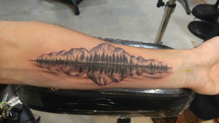Nature tattoo of mountains and trees reflecting on a calm lake, inked on a forearm.