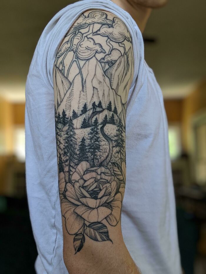 Nature tattoo featuring mountains, trees, and flowers on upper arm, showcasing intricate design.
