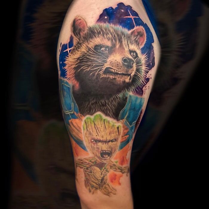 Marvel tattoo featuring a Rocket raccoon character above a small tree-like Groot figure with cosmic background on upper arm.