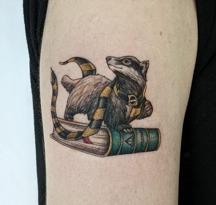 A Hufflepuff-themed Harry Potter tattoo featuring a badger with a scarf atop a book on an arm.
