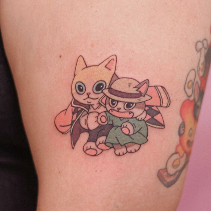Adorable animal-themed tattoo featuring two cartoon cats, one wearing a hat and cloak, on a person's arm.