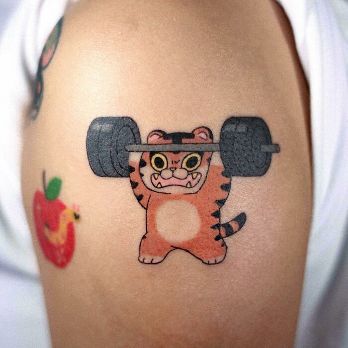 Adorable animal-themed tattoo of a cartoon tiger lifting weights on an arm.
