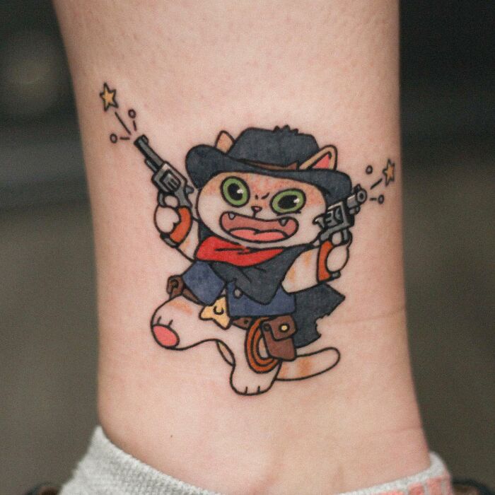 Animal-themed tattoo of a cartoon cowboy cat wearing a hat and holding two guns, with vibrant colors and playful details.