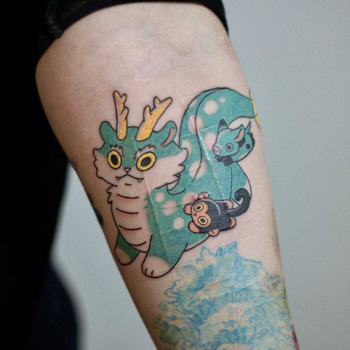 Tattoo of a whimsical green dragon-cat with horns, showcasing adorable animal-themed design on an arm.