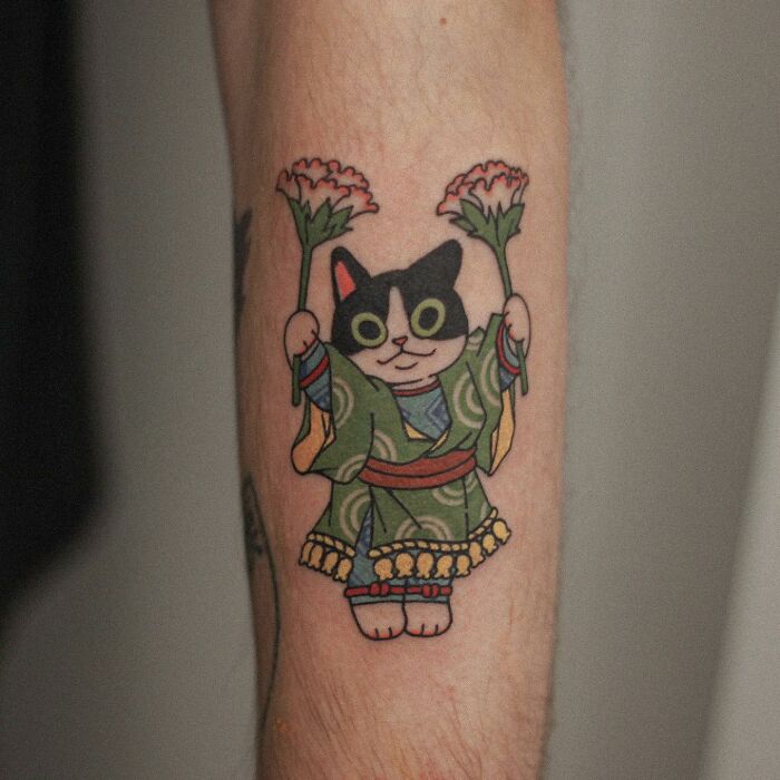 Tattoo of a cat in a green kimono holding flowers, showcasing an adorable animal-themed design.