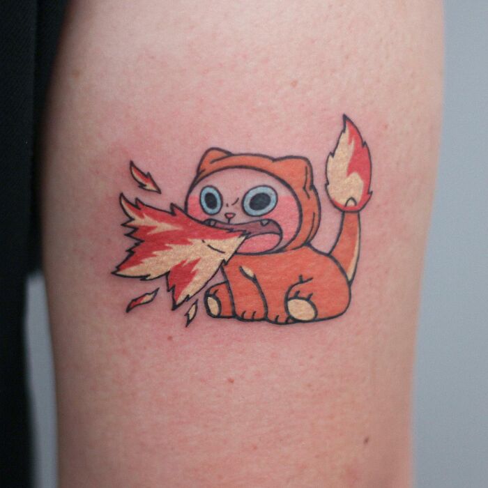 Adorable animal-themed tattoo of a cartoon-like orange cat breathing fire on skin.