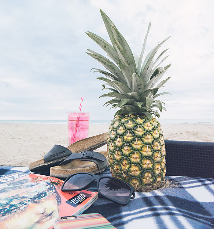 Pineapple, glasses, magazine and flip flops with drink at the beach