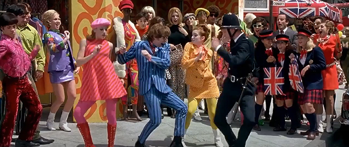 Austin Powers and other characters dressed in color dance in the street