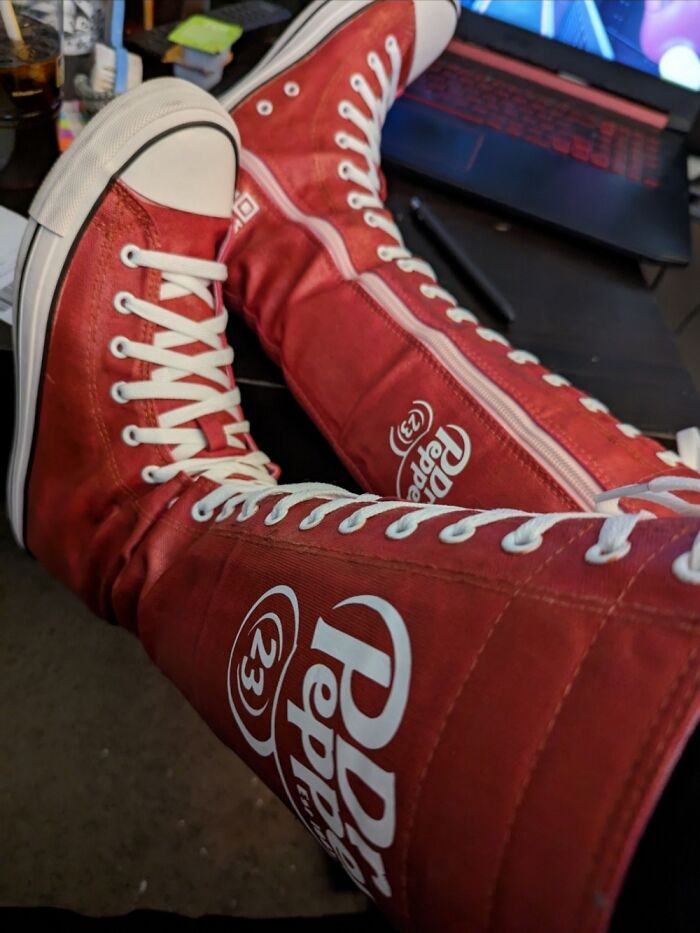 Red high-top shoes with Dr Pepper branding, showcasing unique footwear indoors near a laptop.
