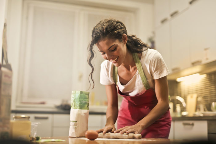 Woman in kitchen baking, smiling in apron, with flour and egg, embodying millennial creativity.