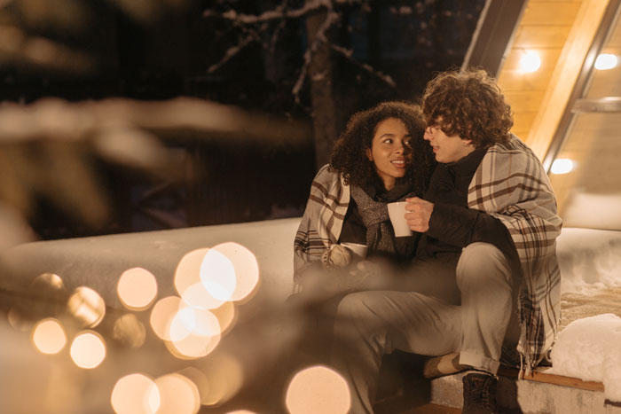 Couple wrapped in blankets outside, enjoying a winter evening, capturing a cozy moment amidst millennial slang charm.