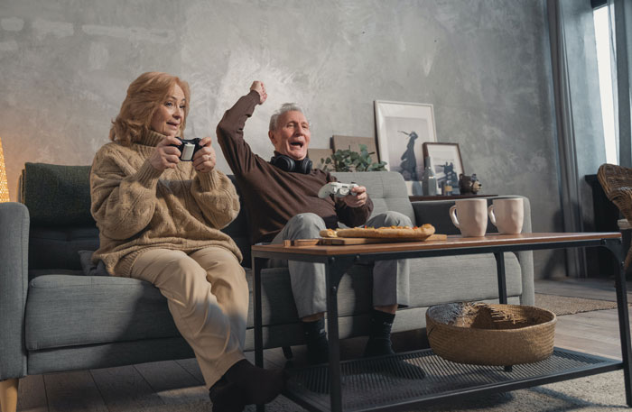 Elderly couple on a couch enjoying video games, embracing millennial slang culture in a cozy living room.