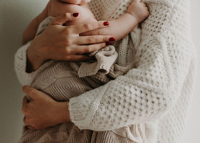 New mom holding baby in soft knit sweater, discussing MIL wanting tattoo of grandkid's footprint. New mom holding baby in soft knit sweater, discussing MIL wanting tattoo of grandkid's footprint.