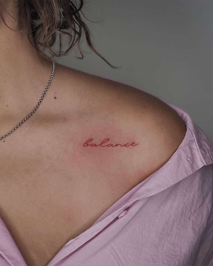 Red ink tattoo reading "balance" on a person’s collarbone, wearing a light pink shirt.