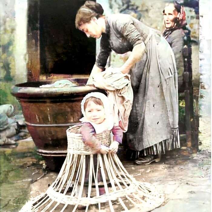 A Baby Learning How To Walk In A Wicker Frame As Mom Does Laundry. 1910s