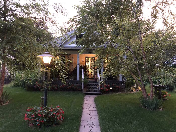 Cozy tiny house design with a green lawn, front porch, and warm lighting surrounded by trees and flowers.