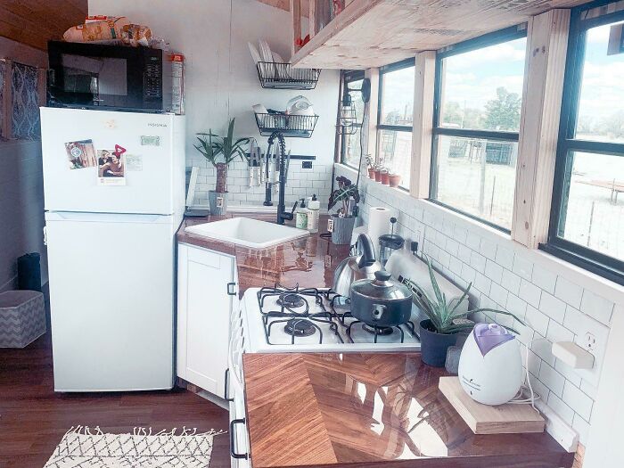 Compact tiny house kitchen featuring white cabinets and brown countertops with stove, sink, and ample natural light.