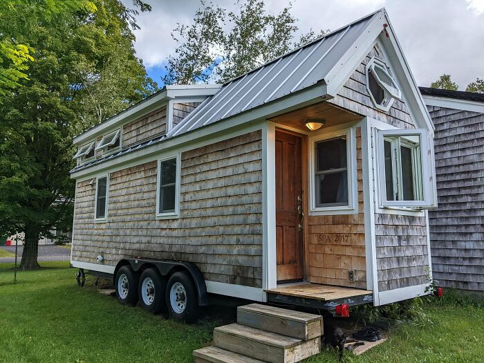 Light grey tiny house on wheels with wooden exterior and metal roof surrounded by green grass and trees.