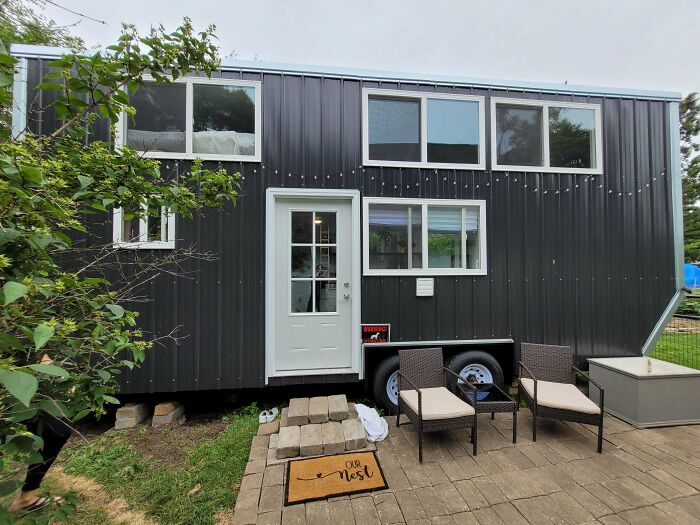 Grey tiny house on wheels with white door and windows, outdoor seating area, and greenery surrounding the compact home design