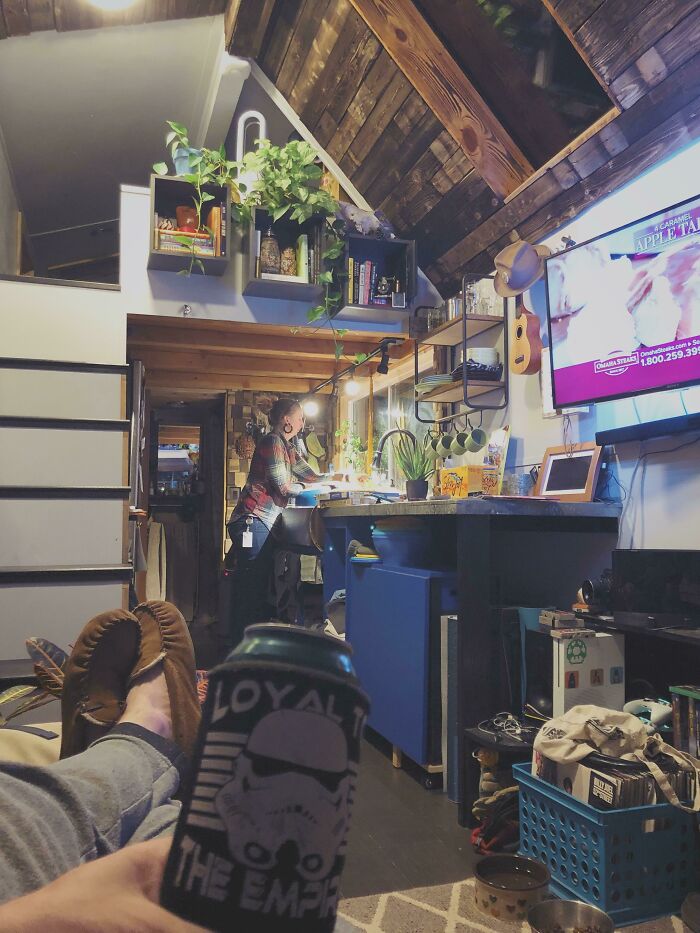 Cozy tiny house interior with a person cooking in the kitchen, plants and a TV visible, showcasing a compact design.
