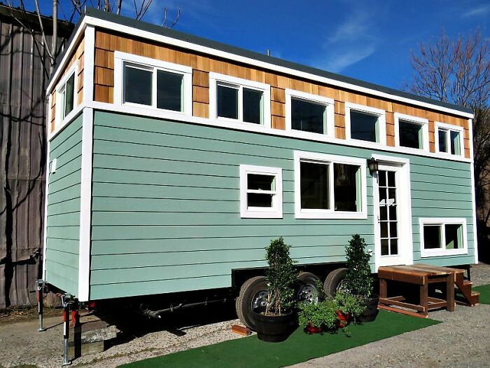 Green and beige small wheel tiny house design with multiple windows and potted plants outside on a sunny day