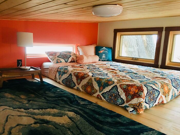 Cozy tiny house bedroom with a patterned bed, orange accent wall, wooden ceiling, and natural light from windows.