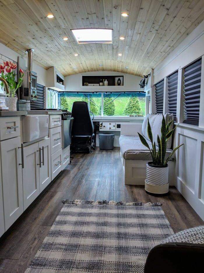 Interior of a tiny house featuring white kitchen cabinets, wooden ceiling, and a cozy living space with a potted plant and rug.
