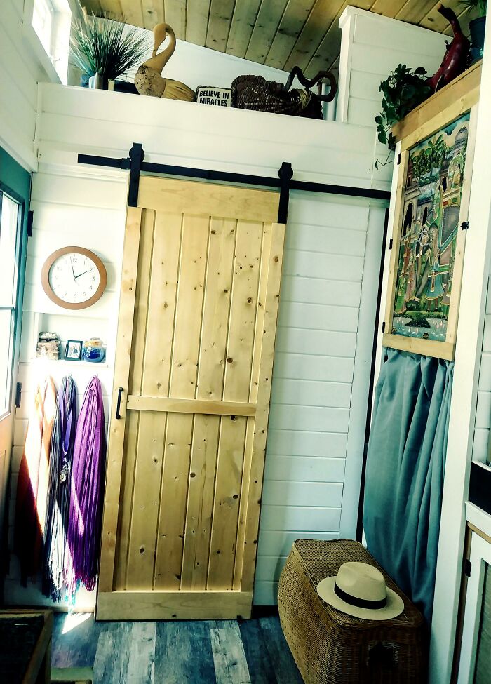Interior view of a tiny house featuring a wooden sliding door, wicker basket with a hat, and colorful scarves hanging by the wall.