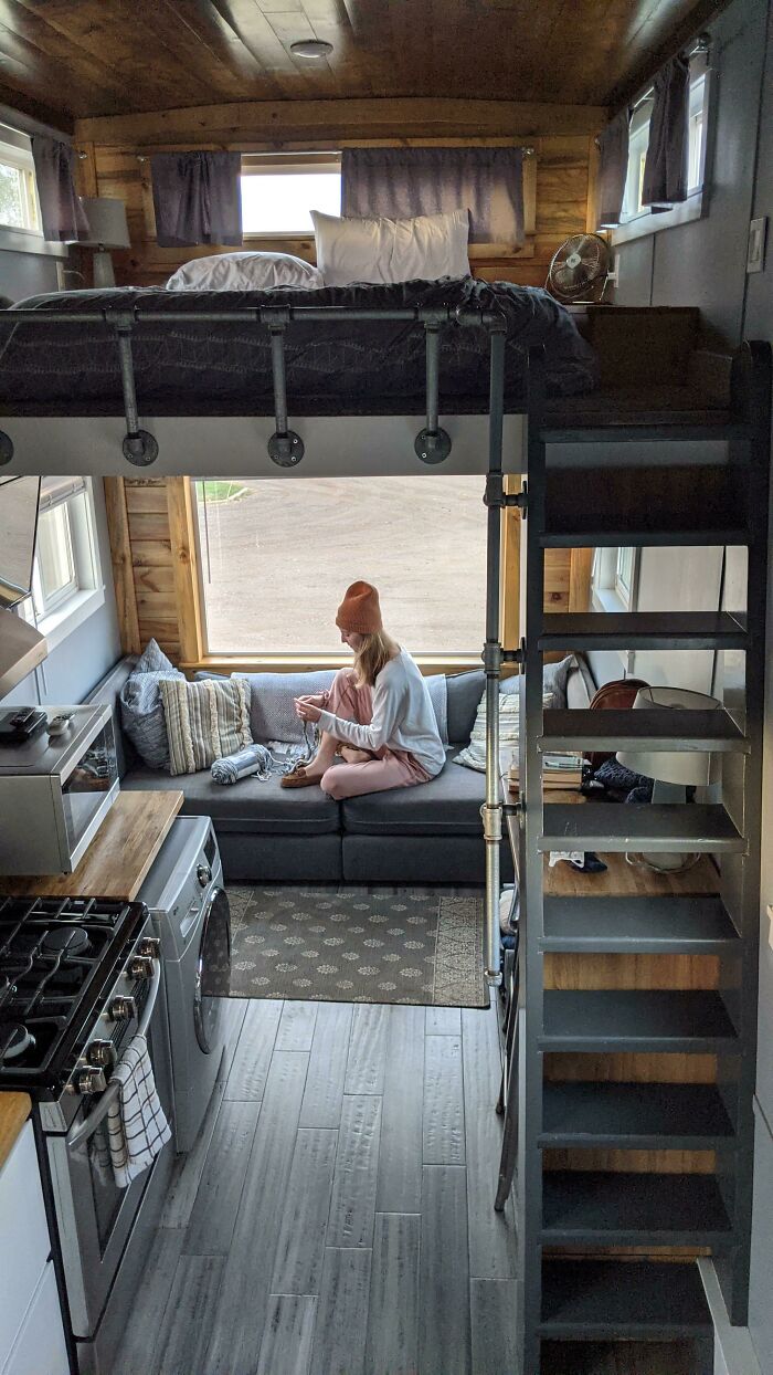 Cozy tiny house interior with lofted bed, woman sitting on sofa, modern kitchen, and sunlight streaming through large window.