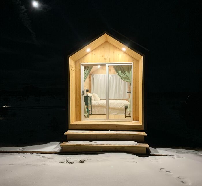 Small wooden tiny house design with large windows, warmly lit interior, and snowy surroundings at night.