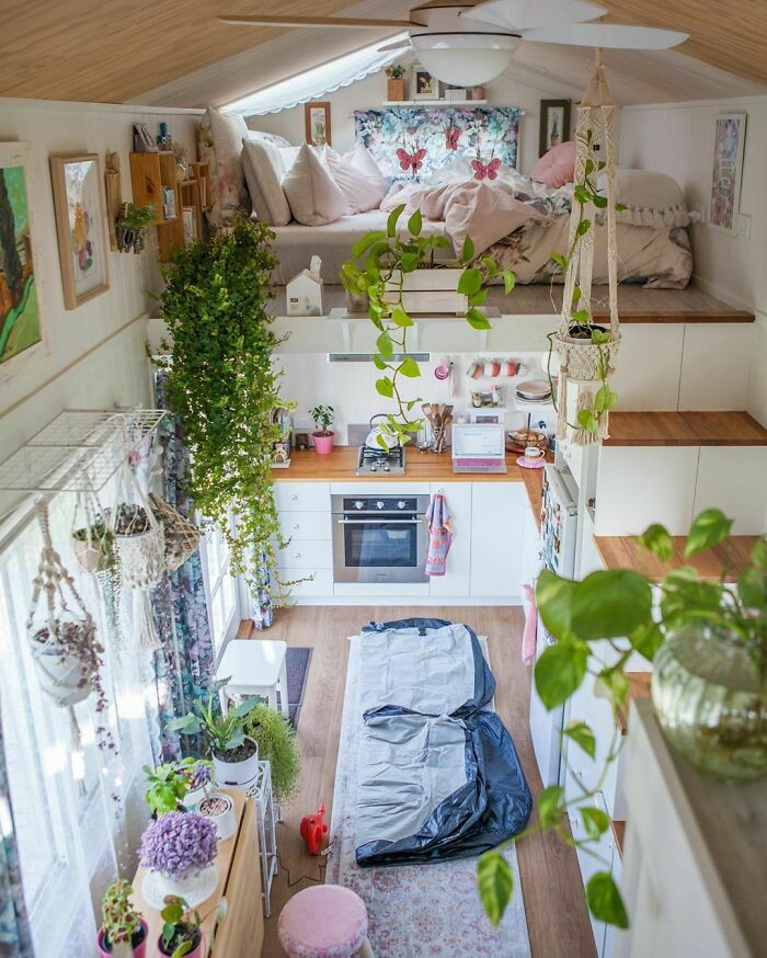 Cozy tiny house filled with lush green plants, featuring a compact kitchen and a sleeping loft with soft bedding.