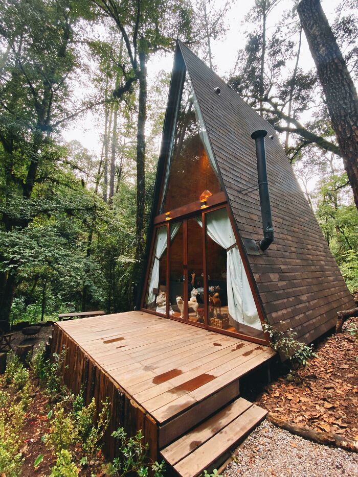 A-frame tiny house design in the woods with large windows, a wooden patio, and cozy interior visible through glass doors.