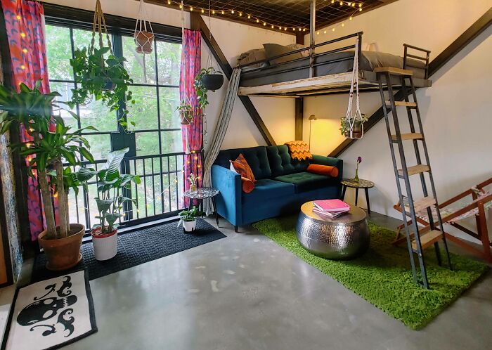 Cozy tiny house interior with a dark blue couch, green rug, loft bed, and hanging plants near large windows.