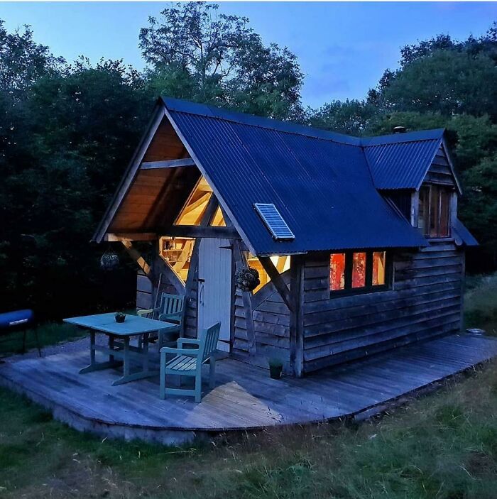 Small wooden tiny house design with patio, illuminated interior, surrounded by trees at dusk in a peaceful setting.