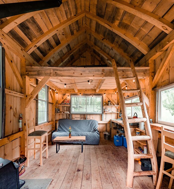 Cozy wooden tiny house interior featuring a grey couch, loft ladder, and natural light from large windows.