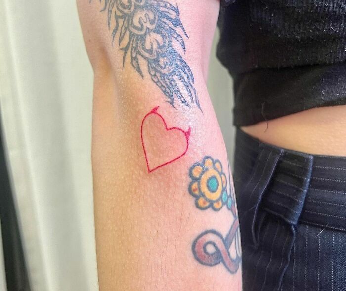 Red ink heart tattoo on arm, alongside colorful floral and black ink designs.