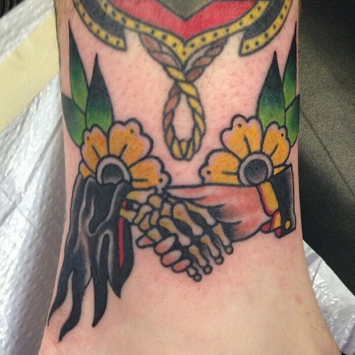 Colorful American traditional tattoo of a skeleton hand shaking a human hand, surrounded by flowers.